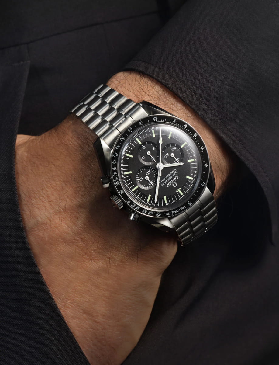 A man’s left wrist wearing a black Omega Speedmaster chronograph with a stainless steel bracelet on a dark sleeve.