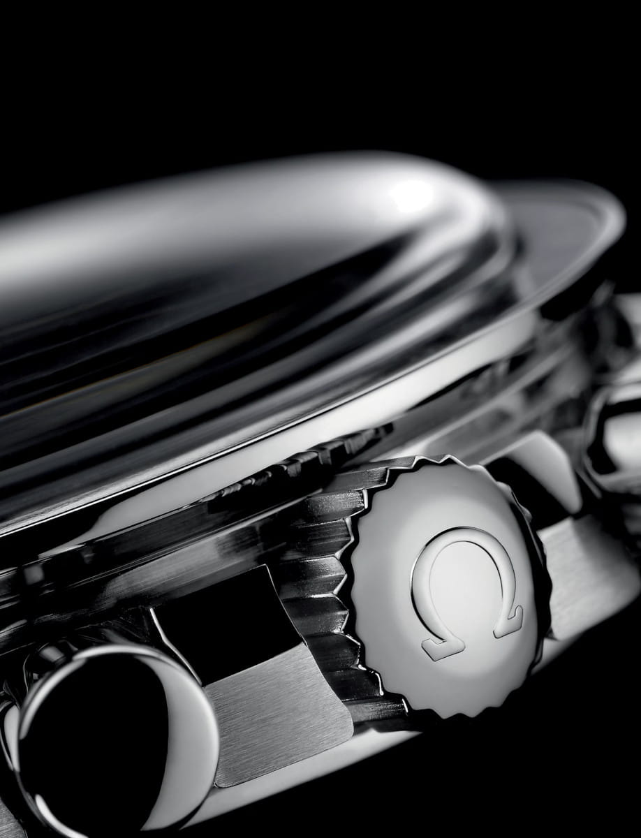Close-up of a stainless steel watch crown with a ridged edge and embossed Omega logo against a dark background.