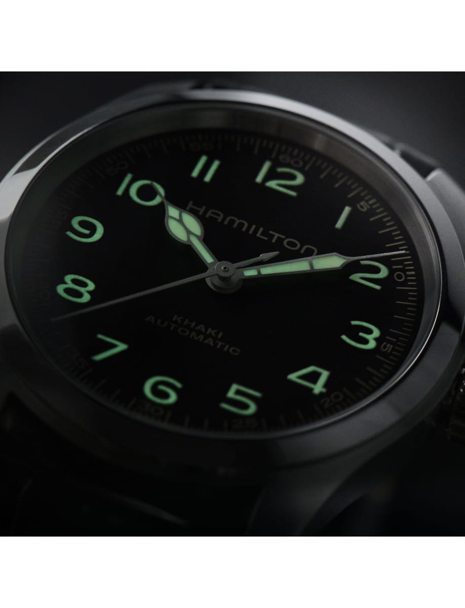 Close-up of a black Hamilton Khaki automatic wristwatch with glowing green numerals and hands in low light