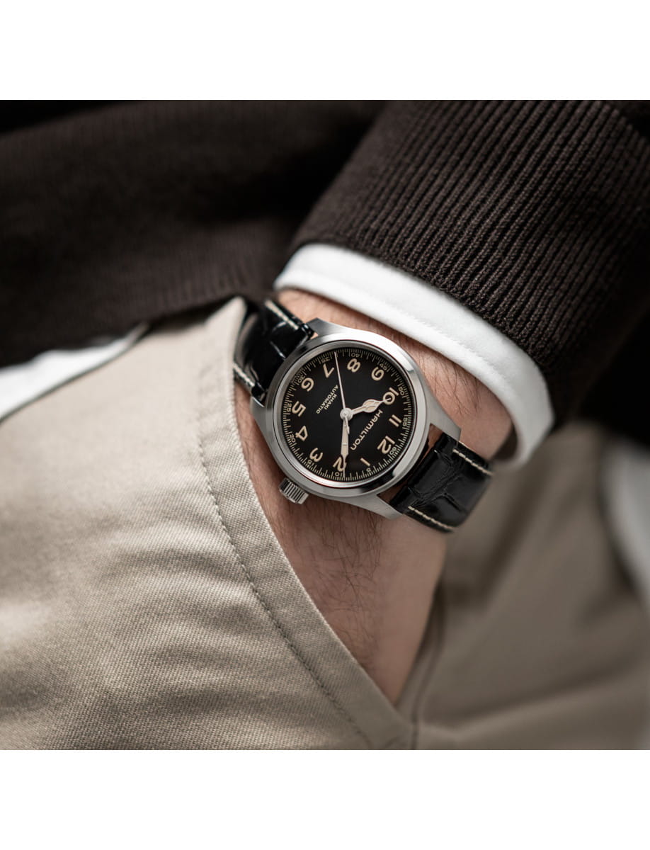 Close-up of a man’s wrist wearing a black-dial leather-strap watch, with a white shirt cuff and beige pants in the background.