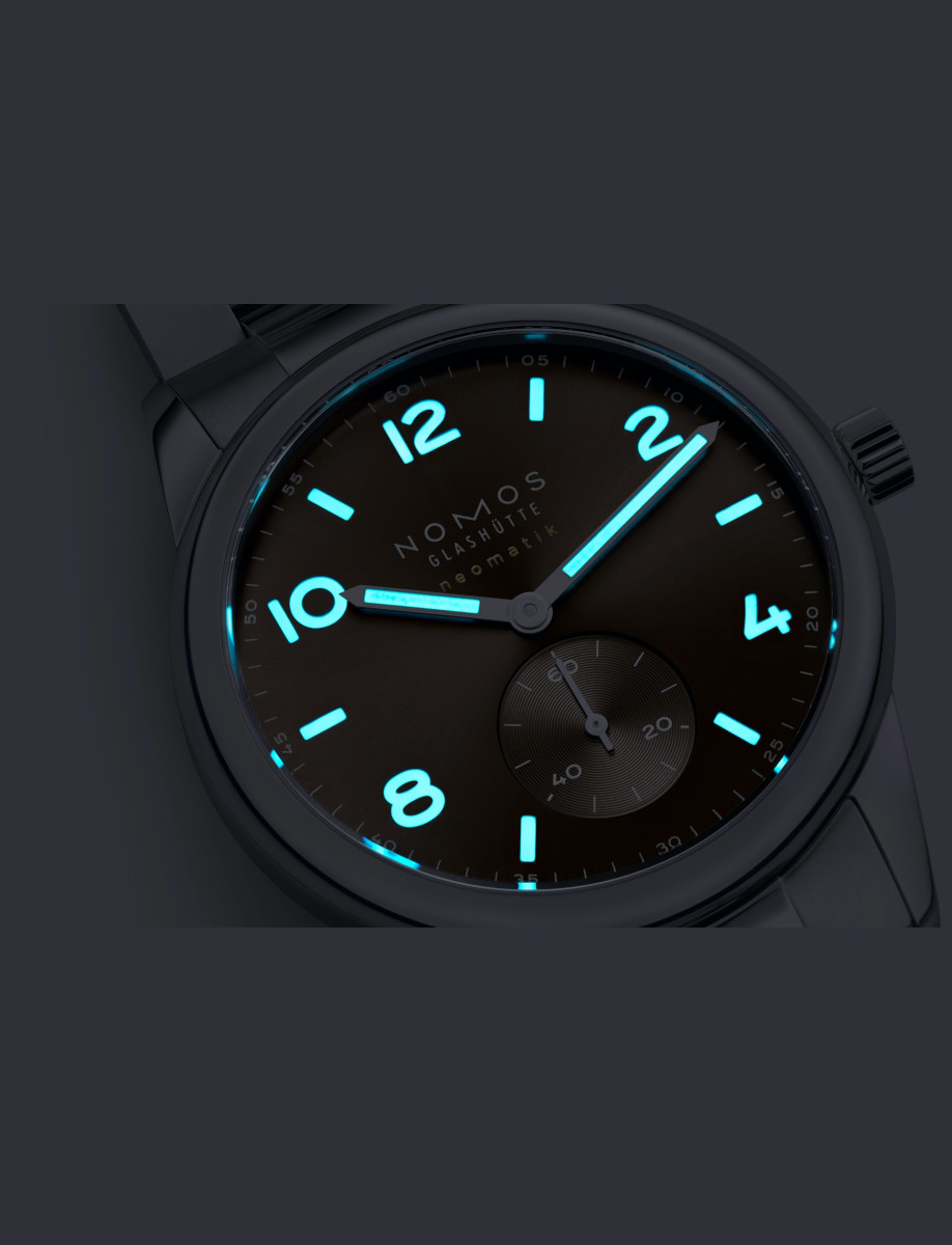 Night view of NOMOS Glashütte wristwatch with stainless steel case and bracelet, and brown dial with luminous hands and hour markers.