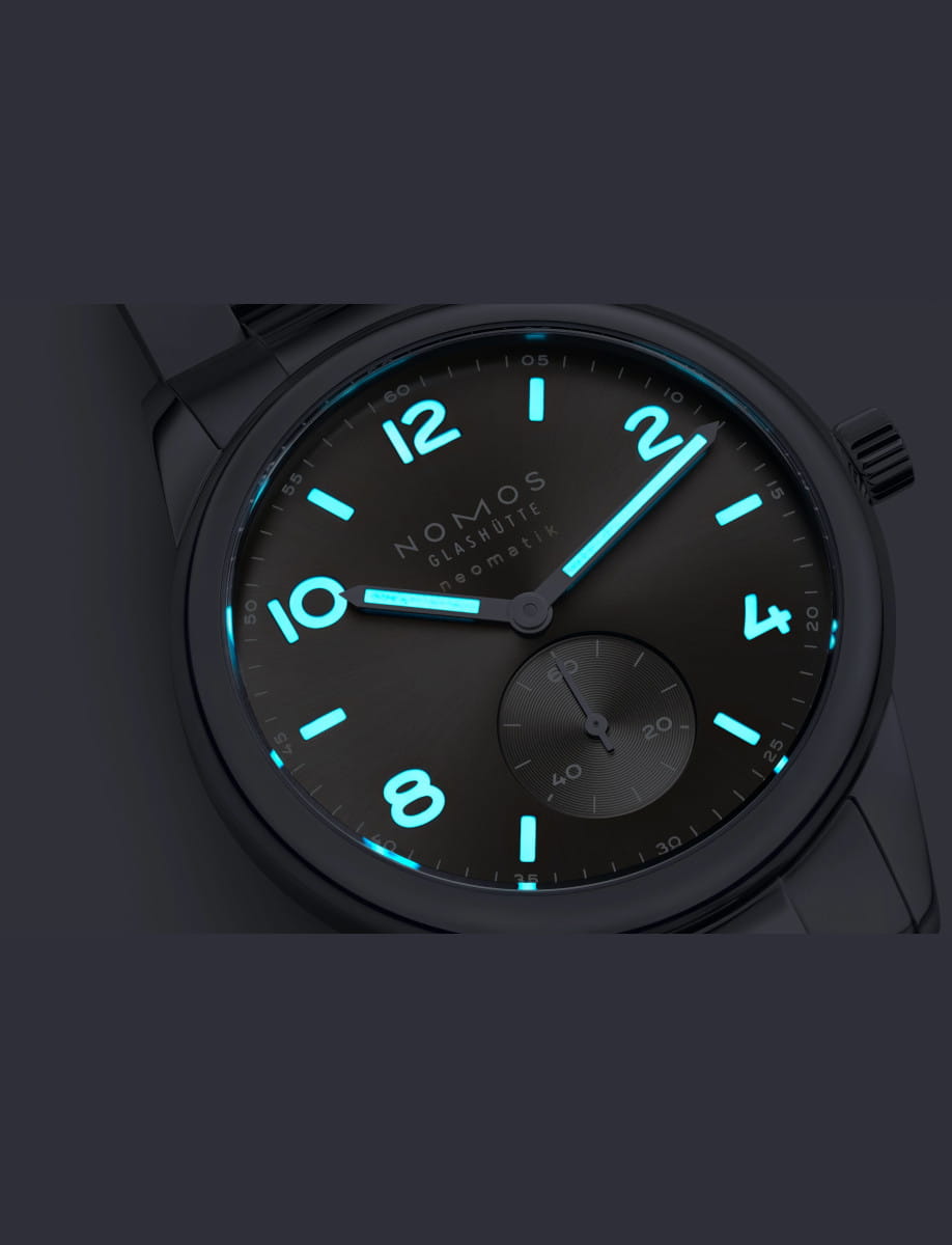 Night view of Nomos Glashutte wristwatch with stainless steel case and bracelet, and light grey dial with luminous hands and hour markers.