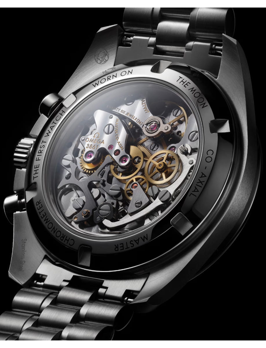 Close-up of a stainless steel watch with a transparent caseback, revealing gold gears and silver components; edge engravings include 'WORN ON THE MOON'.