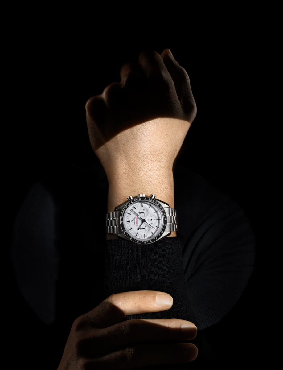 Wrist wearing a stainless steel chronograph watch with a white dial, lit against a dark background.