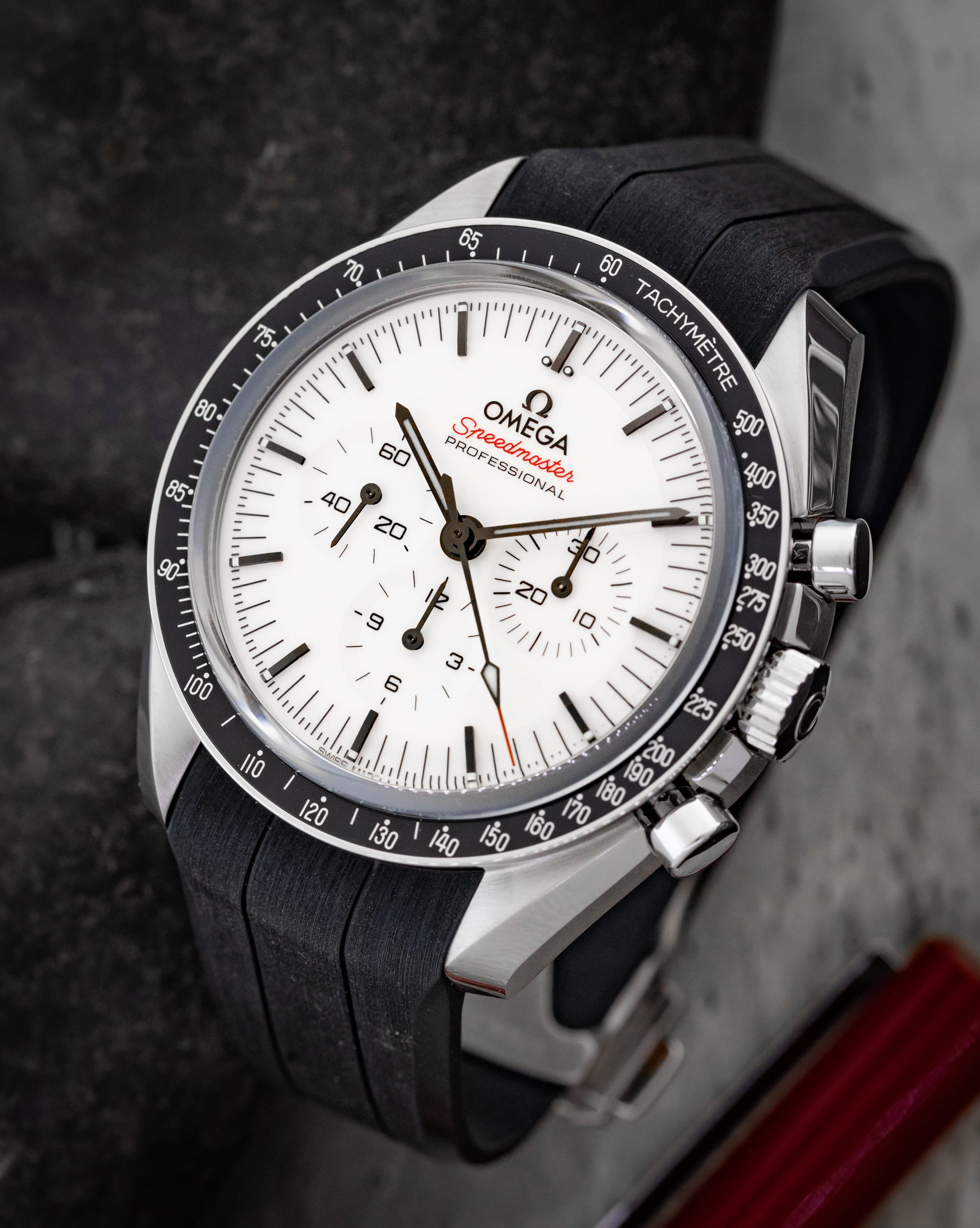 Close-up of an Omega Speedmaster Professional wristwatch showing a white dial and black tachymeter bezel on a dark strap.