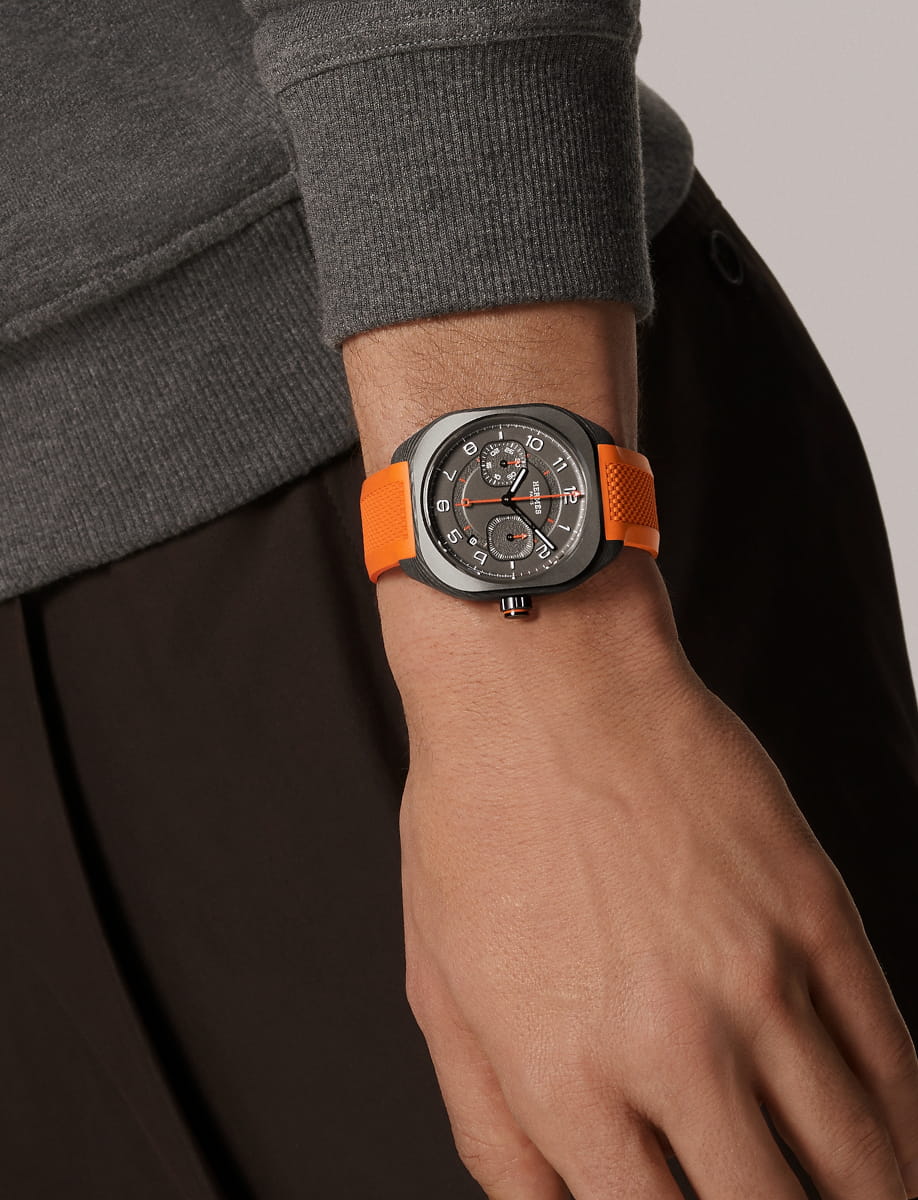 Lifestyle image of Hermes wristwatch with carbon fiber composite case, black gold treated and blackened dial, and orange rubber strap.