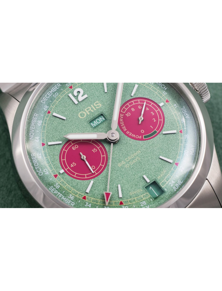 Close-up of a green Oris watch dial with pink subdials, day and month windows, and a power-reserve indicator.
