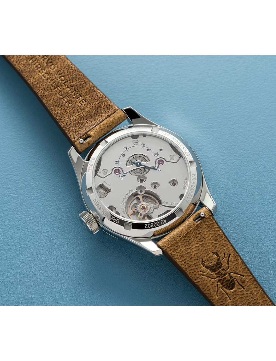 Silver mechanical wristwatch with a transparent case back showing gears and rotor, paired with a brown leather strap on a blue surface.