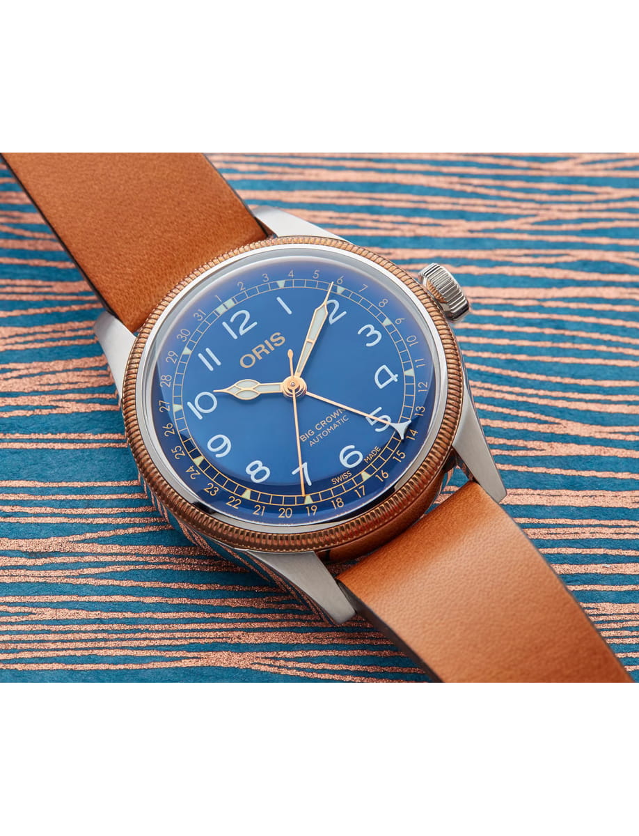 Blue dial wristwatch with gold hands and a brown leather strap resting on a striped surface, showing the ORIS branding and a detailed crown.