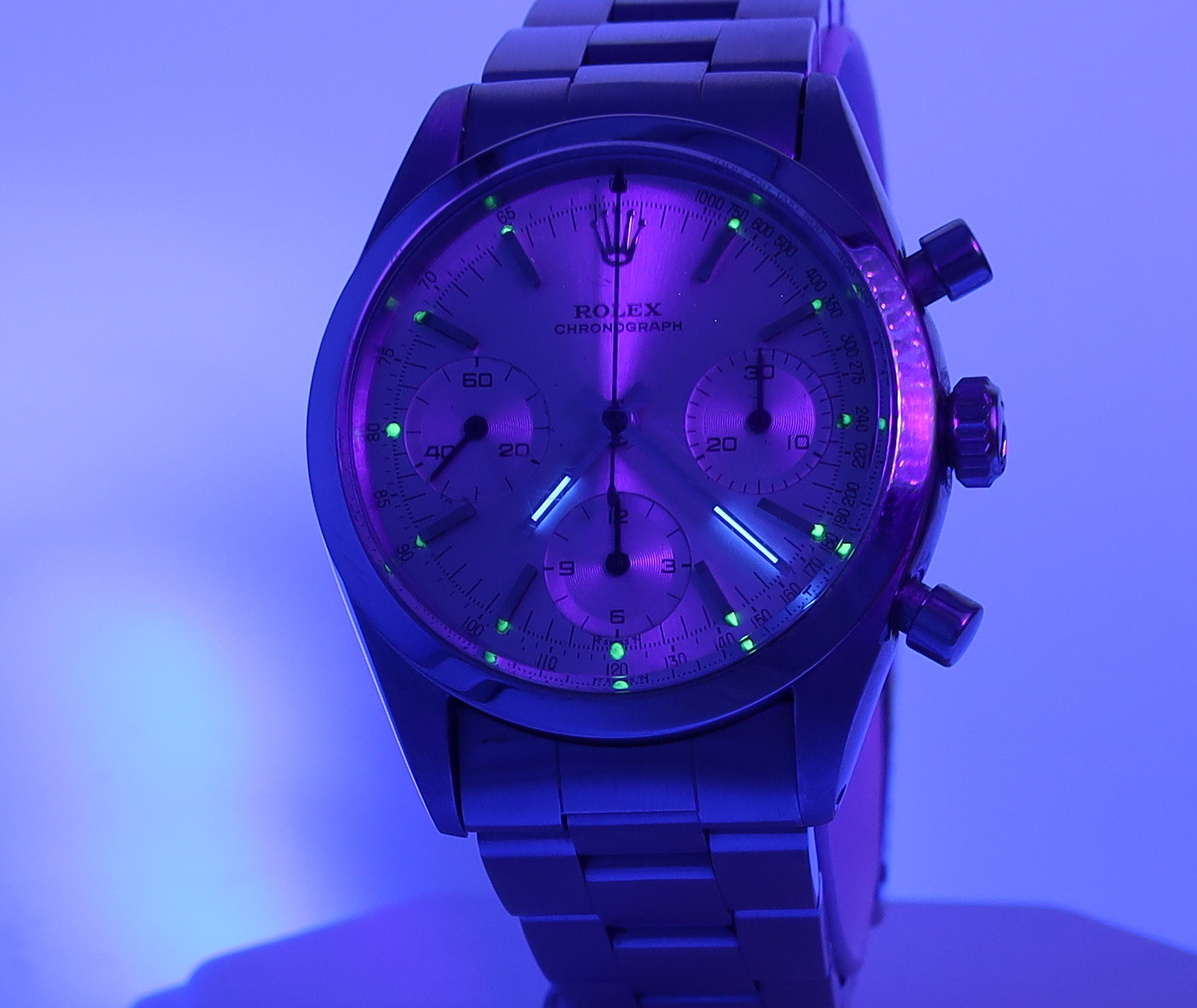 Close-up of a Rolex chronograph wristwatch with a metal bracelet under blue lighting, showing three subdials and glowing markers.