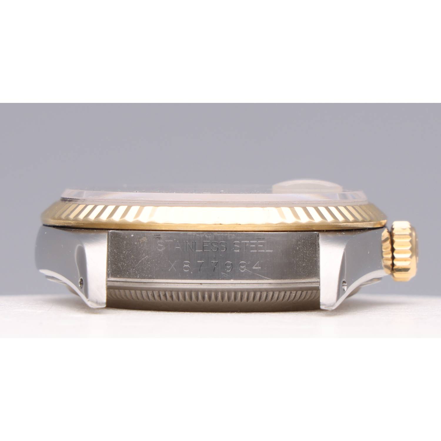 Side view of a two-tone wristwatch with a gold-textured bezel and crown; stainless steel case is engraved with 'STAINLESS STEEL'