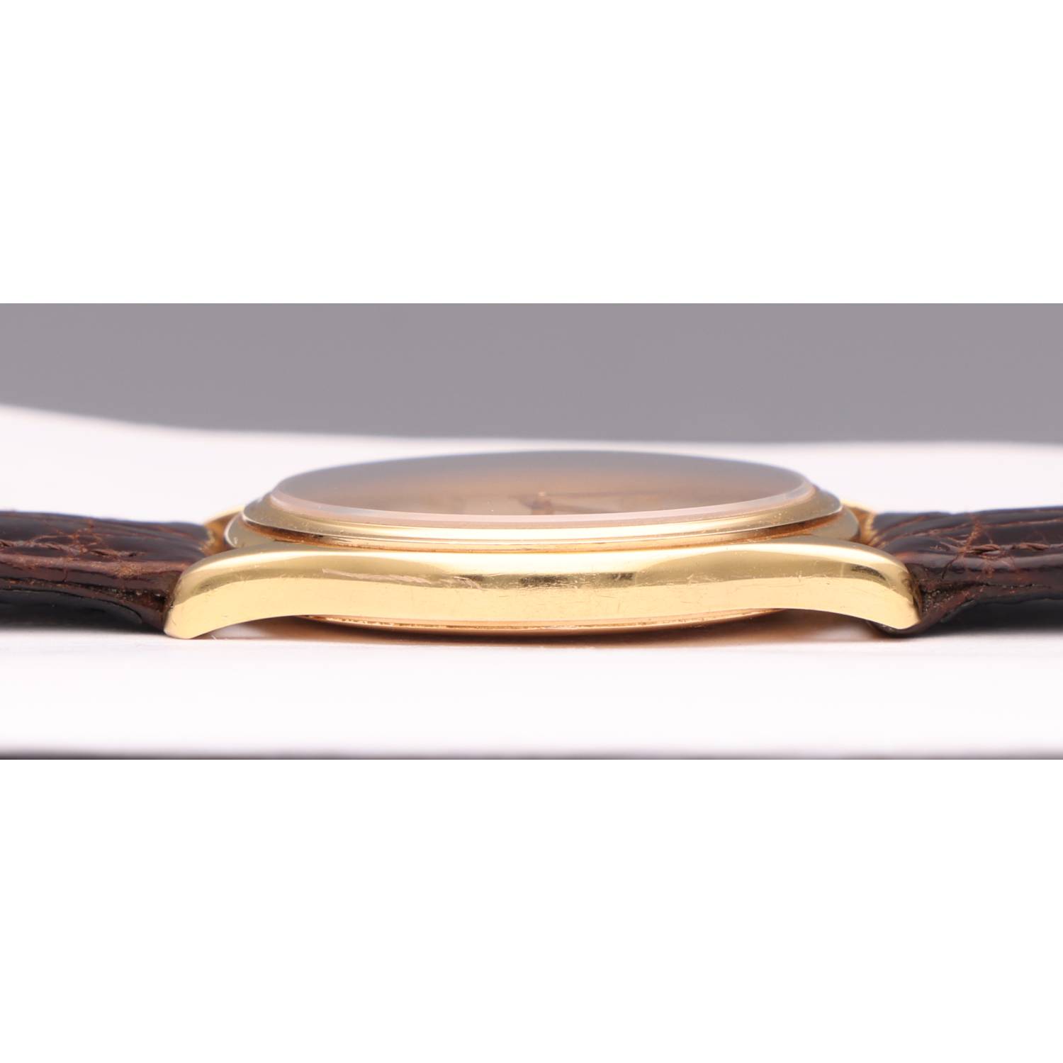 Close-up of a gold wristwatch with a brown leather strap lying on its side on a white surface, seen from the side.