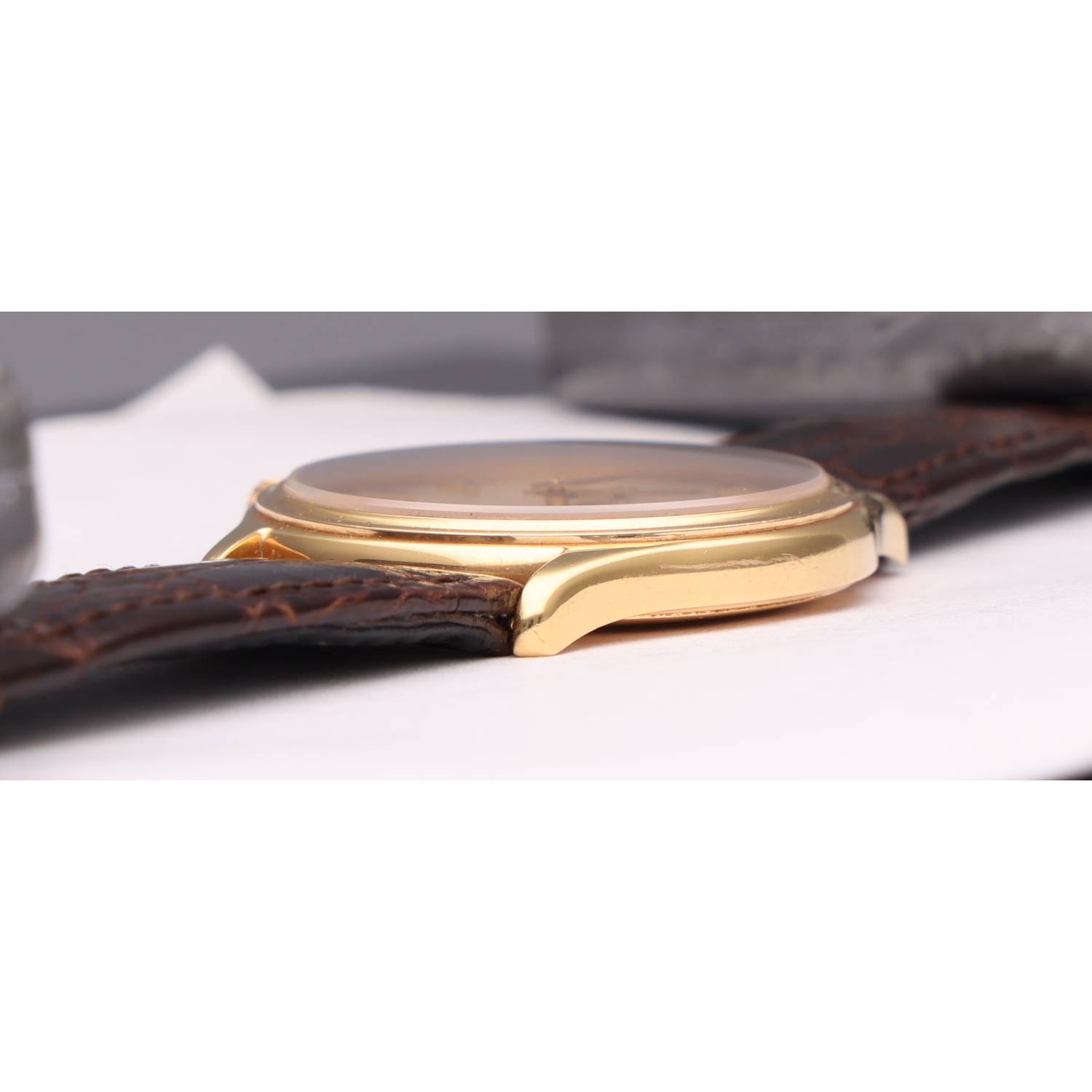 Close-up of a gold-toned wristwatch with a brown leather strap lying on a white surface.