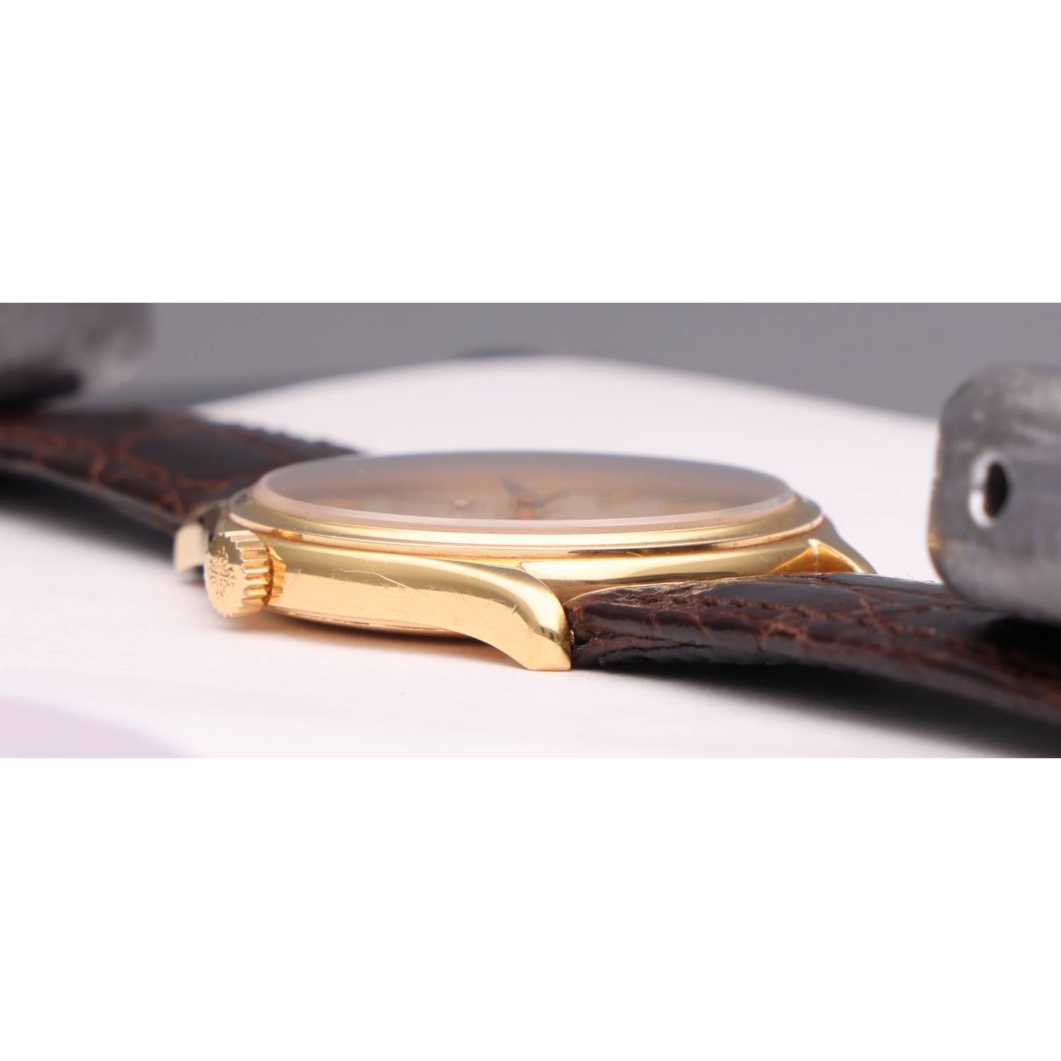 Close-up of a gold wristwatch with a brown leather strap, lying on its side on a white surface.