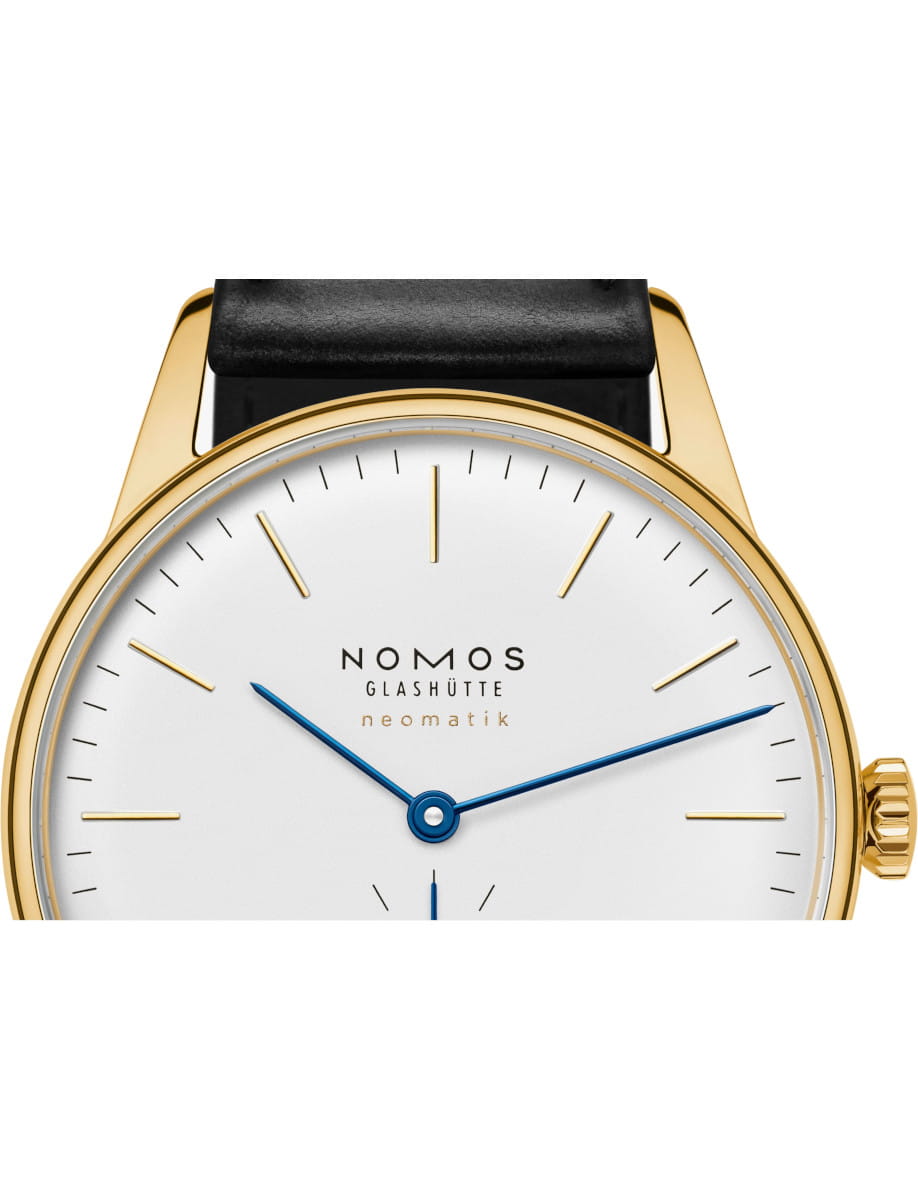 Close-up of a NOMOS Glashütte Neomatik watch with a gold case, white dial, blue hour and minute hands, and a black leather strap edge.