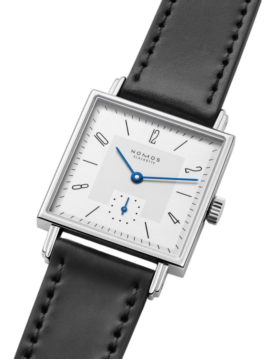 Square-faced NOMOS Glashütte wristwatch with a silver case, white dial, blue hands, and a black stitched leather strap