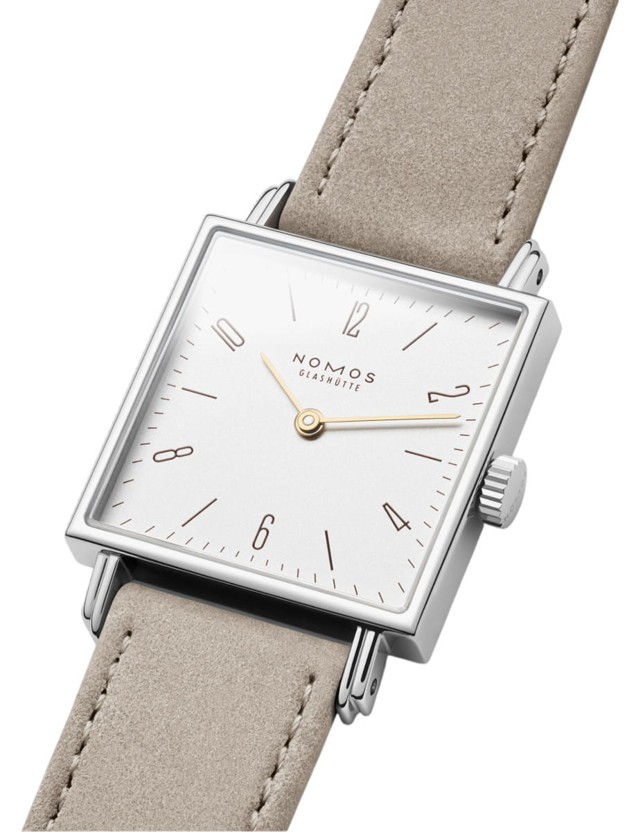 Square silver wristwatch with a white dial, gold hands, and a beige suede strap; Nomos Glashütte logo visible on the face.