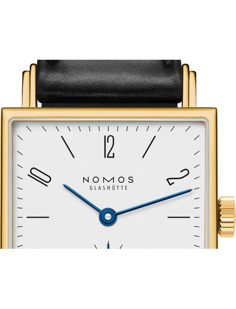 Close-up of a NOMOS Glashütte wristwatch with a gold square case, black leather strap, white dial, and blue hands showing around 12 o'clock.