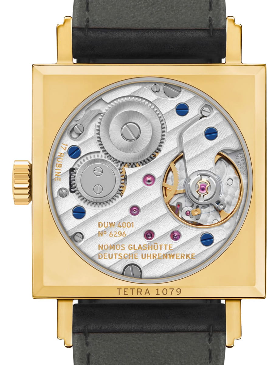 Gold square watch case with visible mechanical movement and blue screws, leather strap, markings 'TETRA 1079' and 'NOMOS GLASHÜTTE DEUTSCHE URENWERKE' on the plate