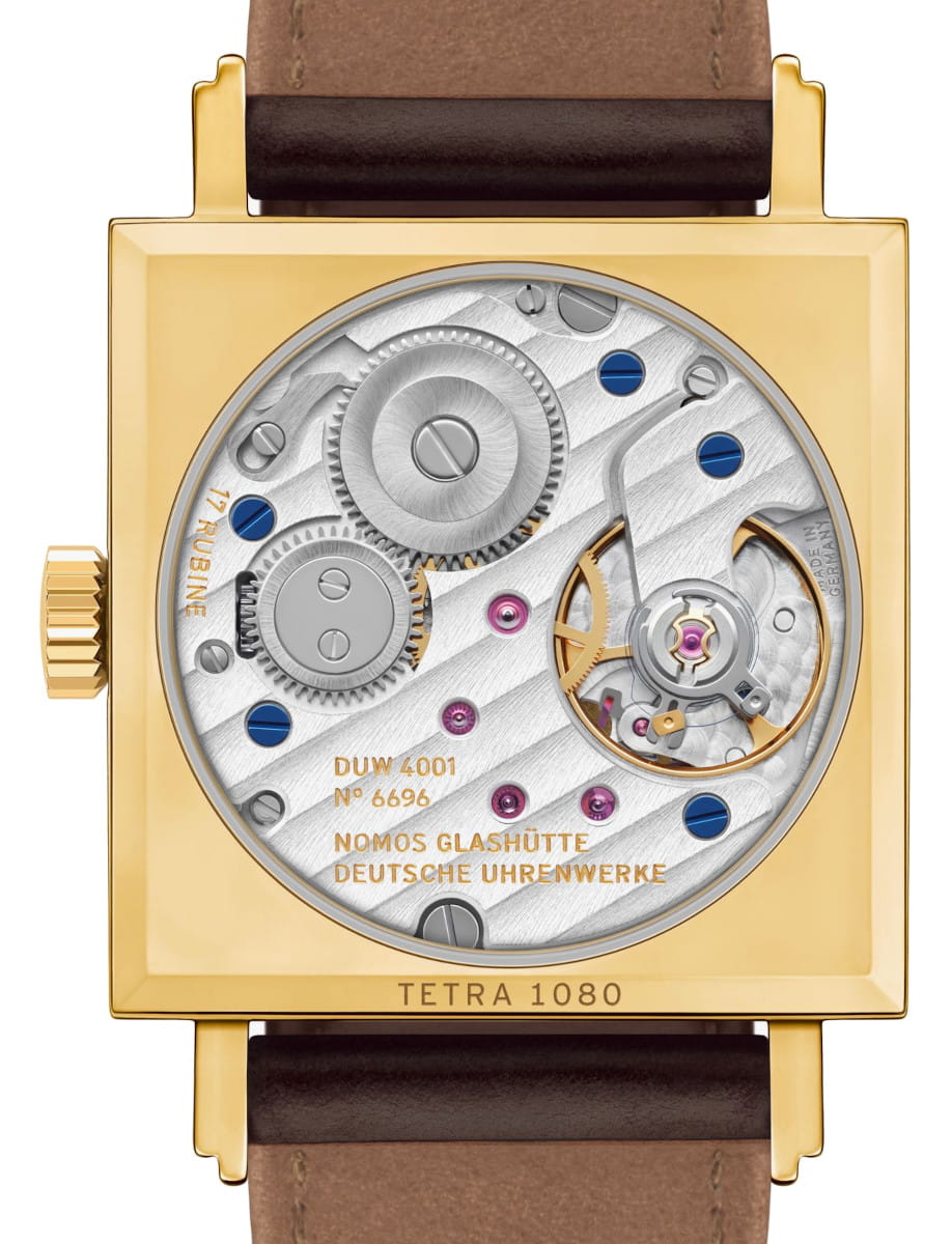 Gold square wristwatch with an exposed mechanical movement, showing gears and blue screws; engravings include NOMOS GLASHÜTTE DEUTSCHE UHRENWERKE and TETRA 1080 on the movement. Brown leather strap partially visible.
