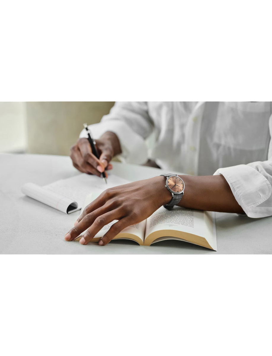 Person writing in a notebook at a desk, with an open book and a wristwatch visible on the arm