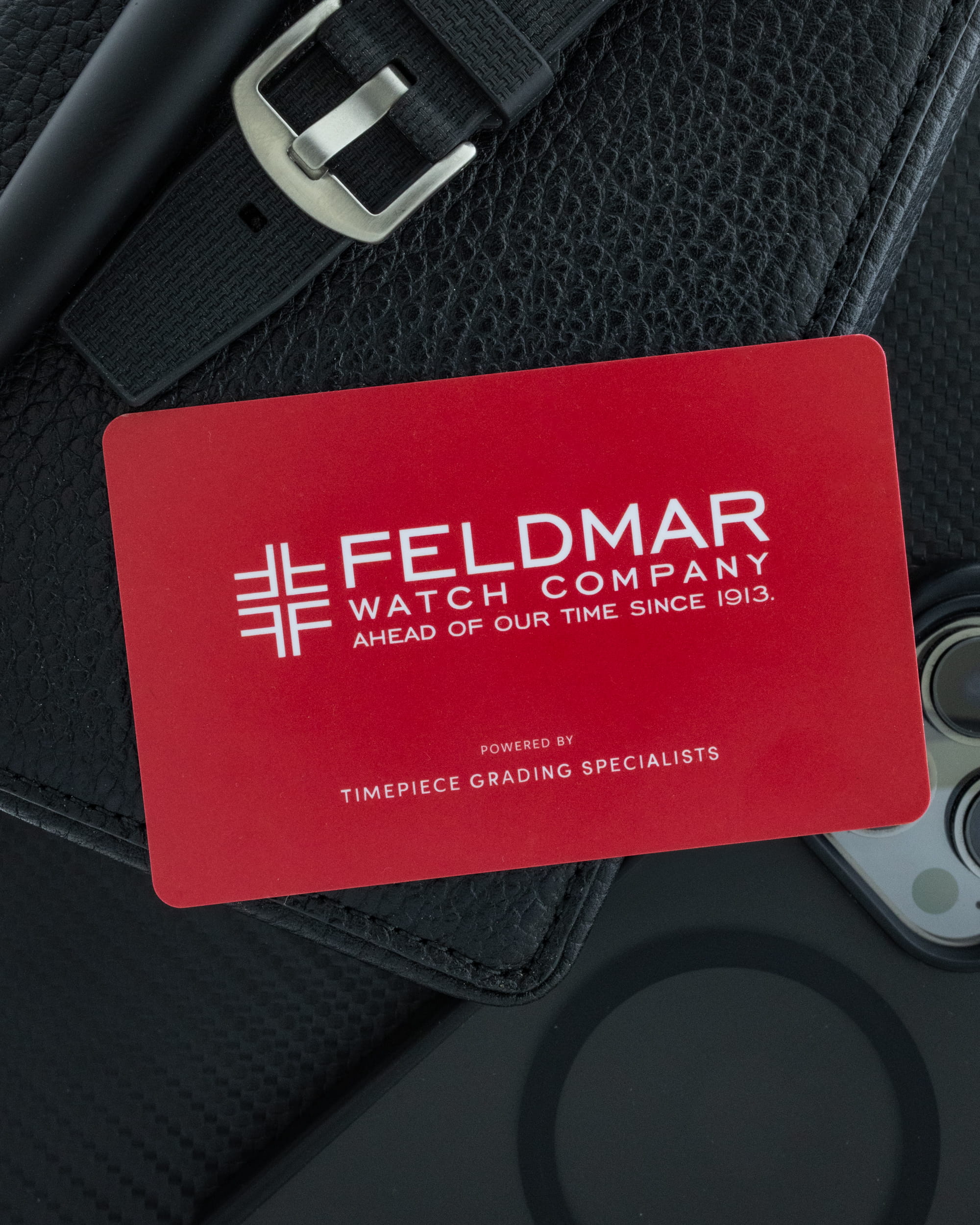 Close-up of a red card with white branding resting on a textured black surface beside a black belt buckle, suggesting a product display.