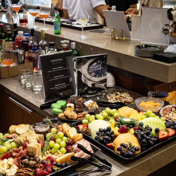 Buffet counter with an assortment of fruits, cheeses, pastries, and drinks, while people chat in the background at a bar area.