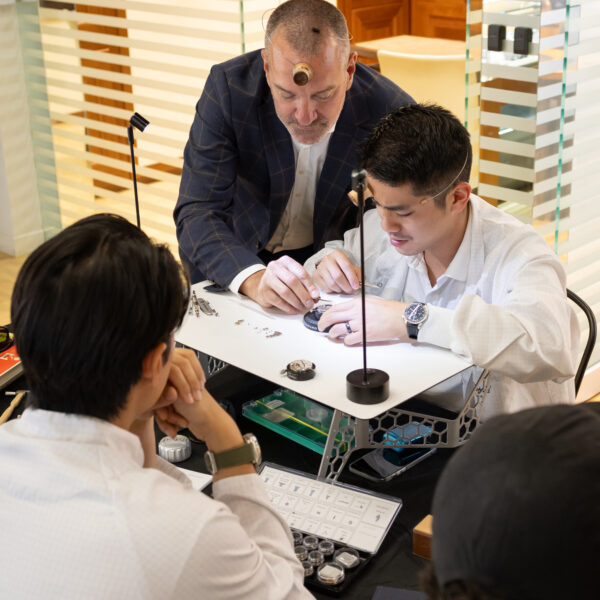 Group of men in a watchmaking workshop examining watches with magnifying stands and small tools on a white table