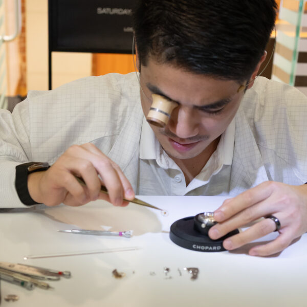 Watchmaker at a clean white workbench assembling tiny watch parts with precision tools, using a loupe to inspect components.