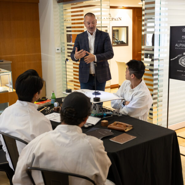 Man in a checked blazer stands and explains to a group of people in white coats seated around a table in a watchmaking workshop showroom.