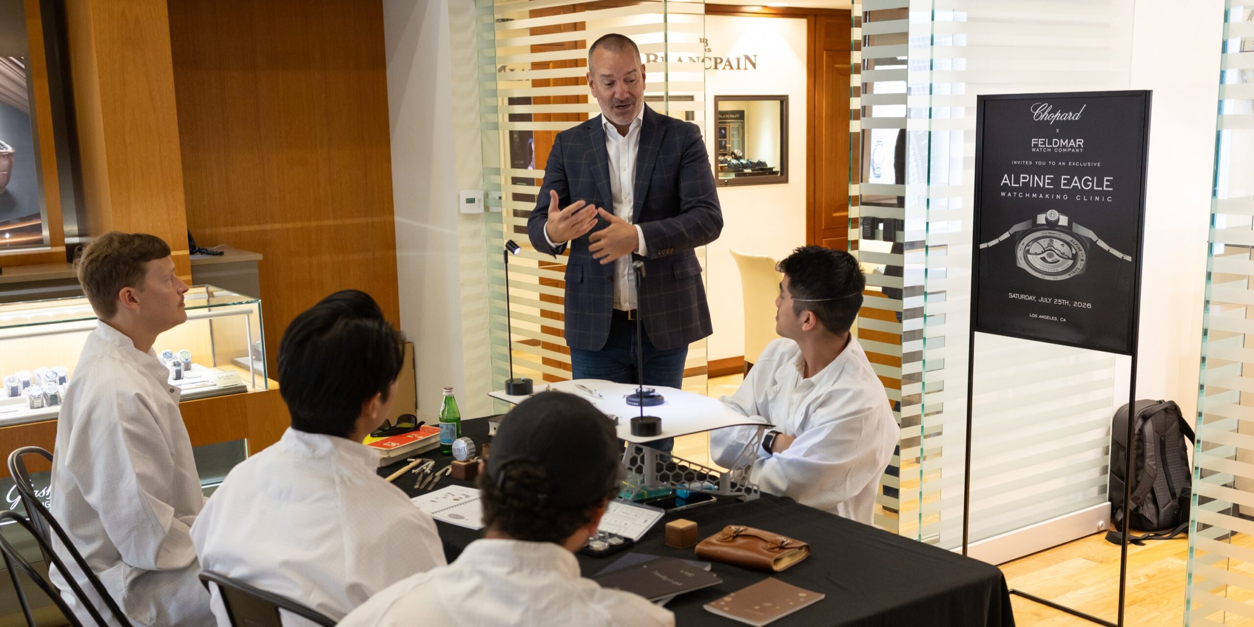 Man in a navy blazer speaks to a seated group in white coats at a luxury watch showroom, with display cases nearby.