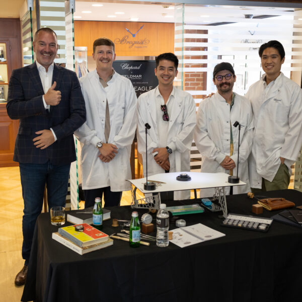 Five men pose for a photo at a jewelry workshop or showroom; one in a dark blazer gives a thumbs‑up while four others wear white coats and smile.