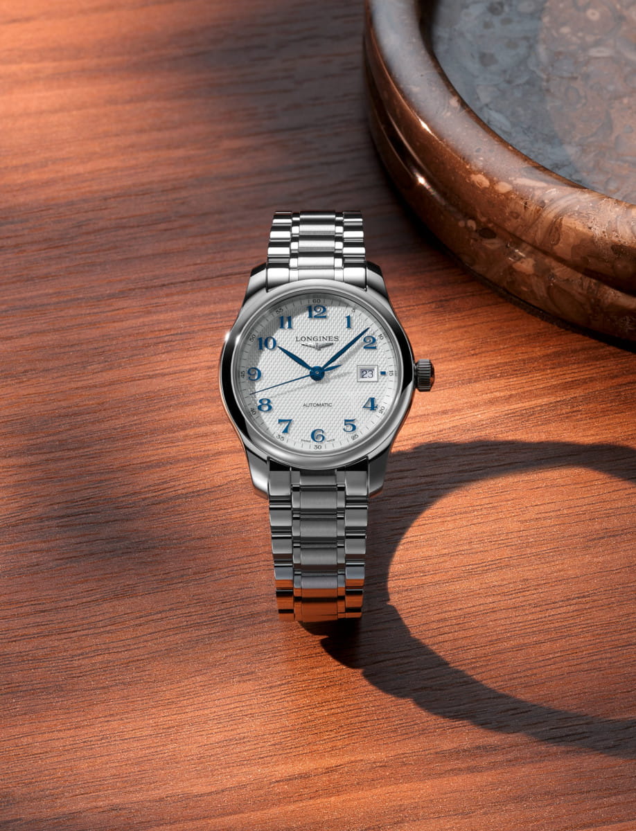 Steel Longines wristwatch with a silver dial and blue numerals resting on a wooden table next to a ceramic bowl edge.