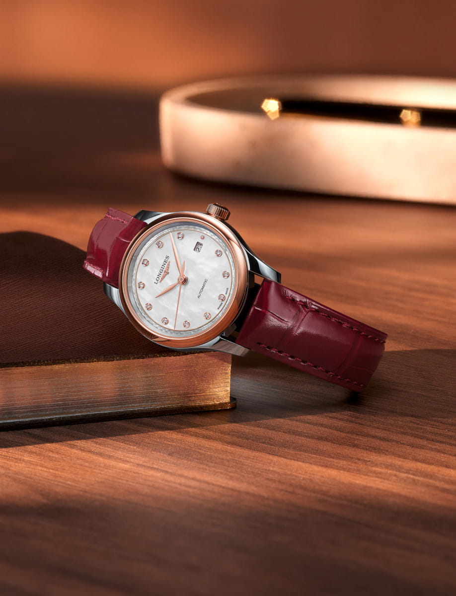 Rose gold Longines wristwatch with a white mother-of-pearl dial and diamond hour markers on a bordeaux leather strap, resting on a wooden surface.