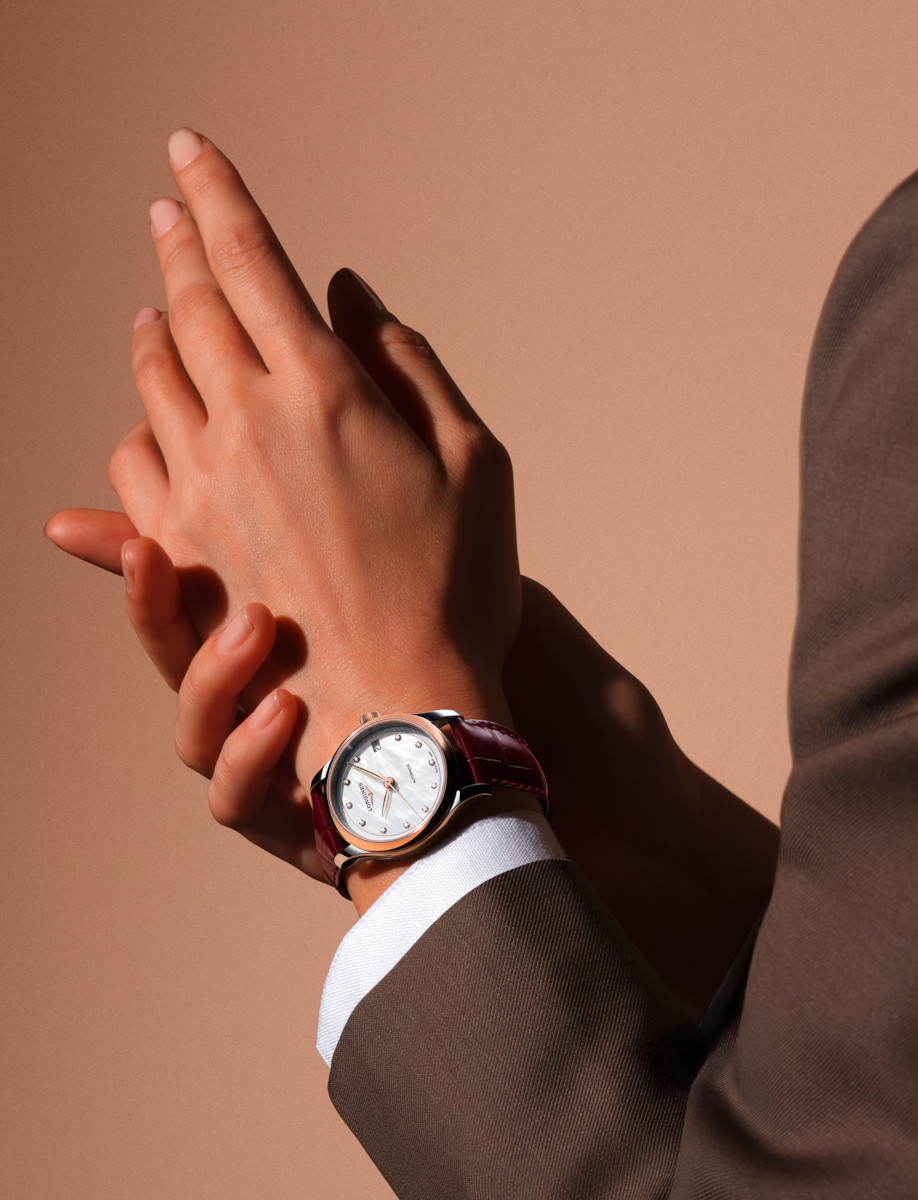 Lifestyle image of Longines automatic wristwatch with a stainless steel case and rose-gold bezel, mother-of-pearl dial, diamond hour markers, and bordeaux leather strap (date window at 3 o'clock).