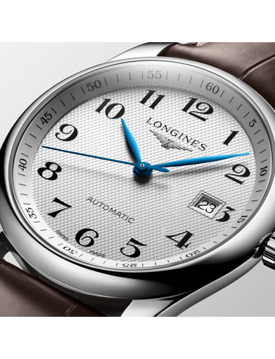Close-up of a silver Longines watch with blue hands and a date display set to 23 on a white textured dial, brown strap visible behind the case.