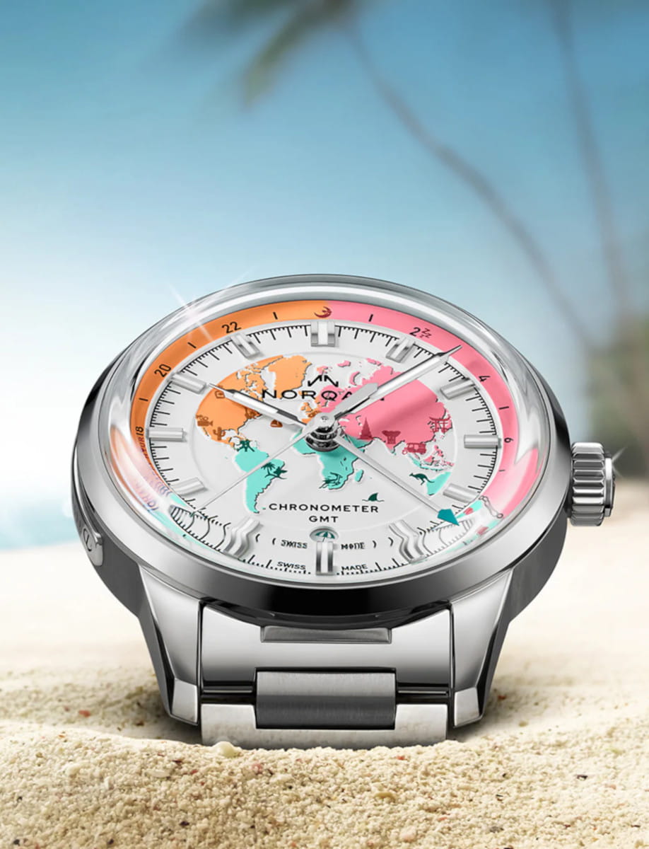 Norqain stainless steel wristwatch with a world map dial and GMT scale resting on sandy beach.