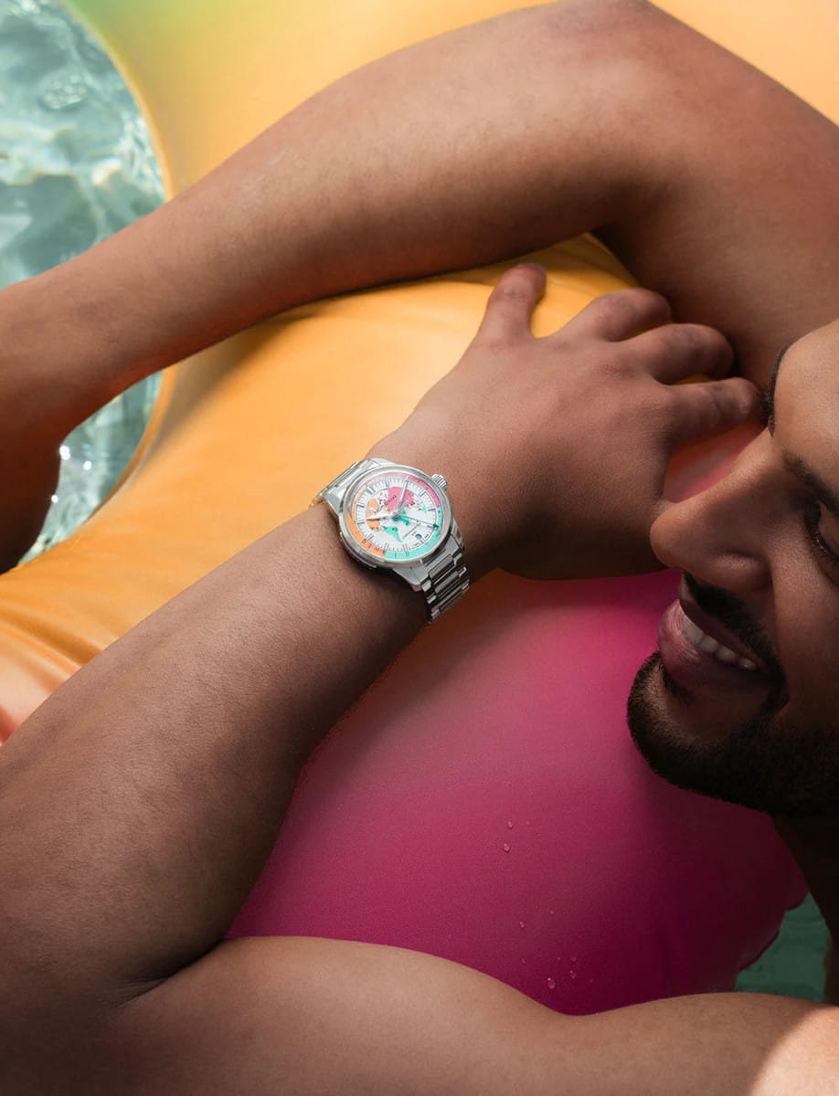 Close-up of a man lounging on a multicolored inflatable pool float, wearing a Norqain stainless steel wristwatch with a white and pastel dial.
