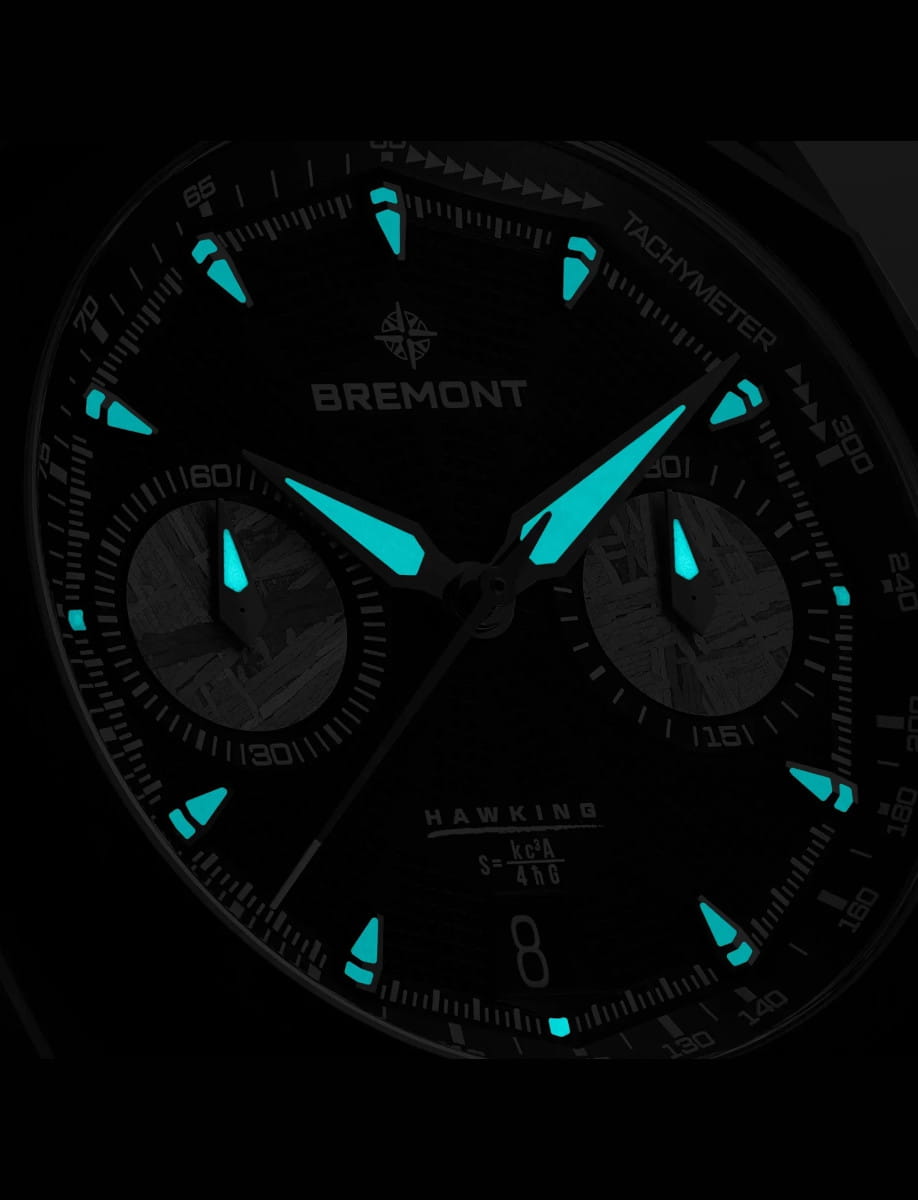 Close-up of a Bremont Hawking watch dial with turquoise luminescent hands and markers at night.