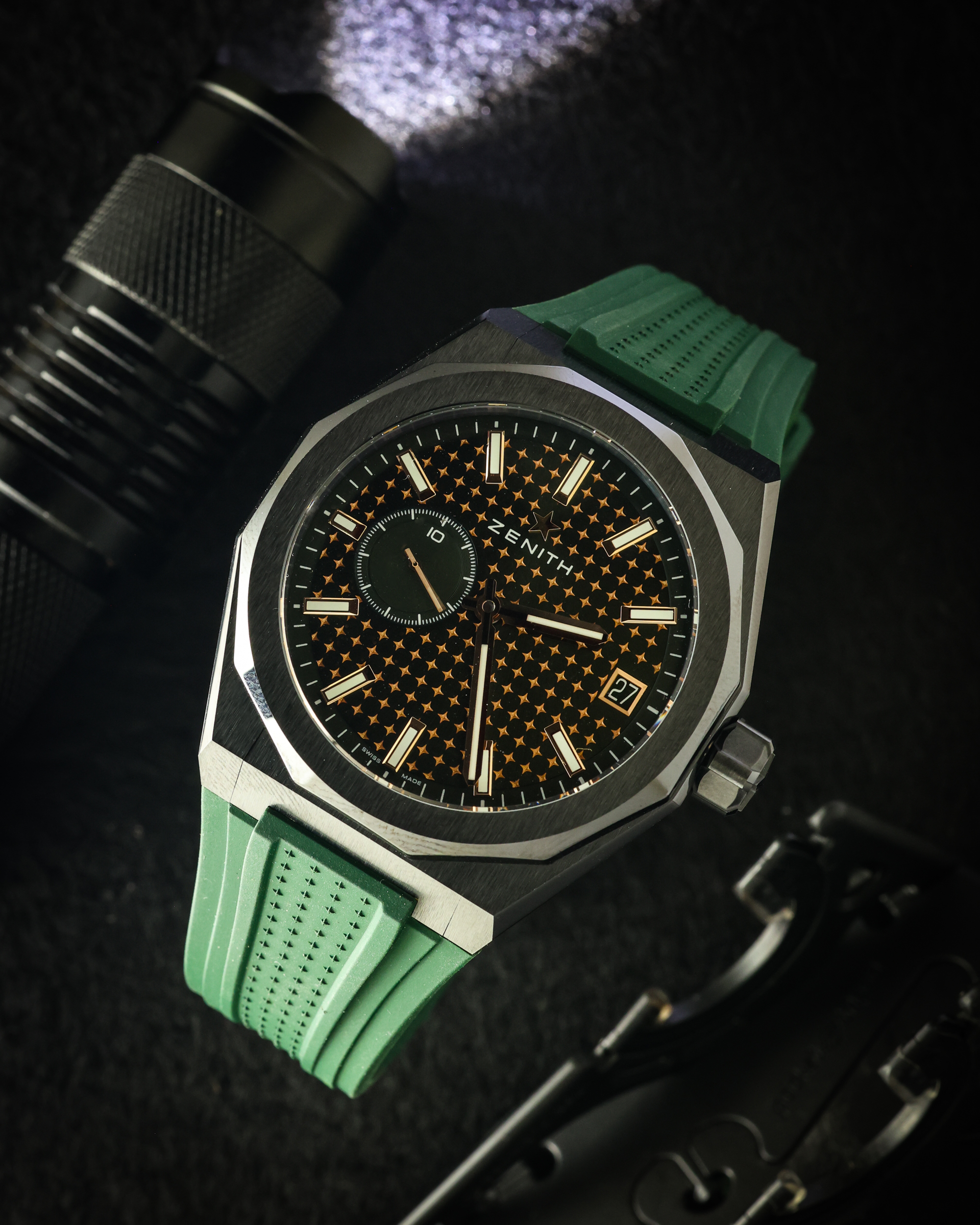 Close-up of a Zenith wristwatch with a green strap, black dial with an orange star pattern, gold-tone indices, and a date window at 3 o'clock.