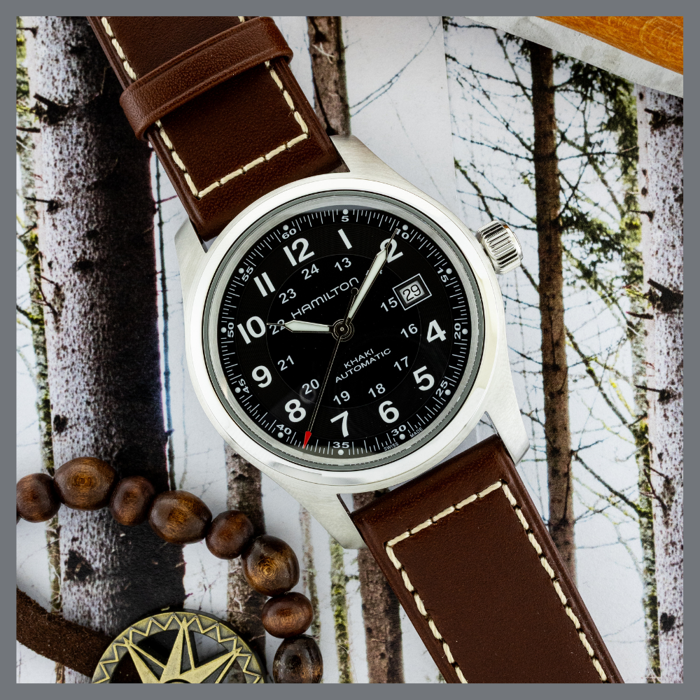 america250 8 | Feldmar Watch Co. Hamilton Khaki automatic wristwatch with a brown stitched leather strap on a nature-themed backdrop, alongside a beaded bracelet.
