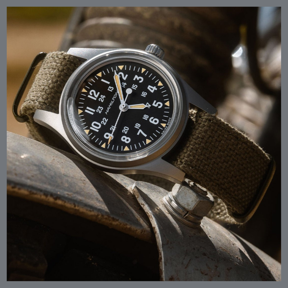 america250 | Feldmar Watch Co. Military-style wristwatch with a black dial, white numerals, and tan markers on a rugged canvas strap resting on a metal surface.