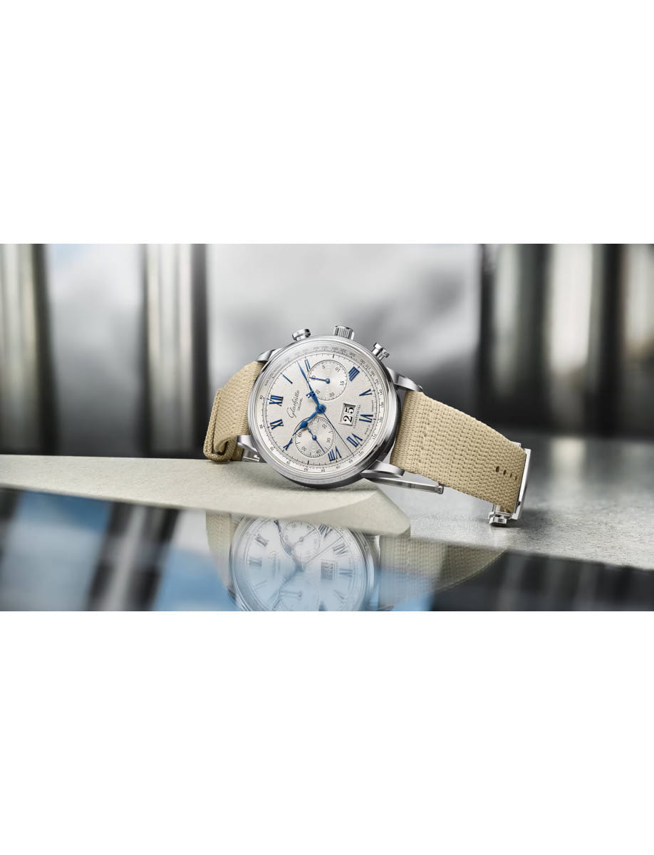 Silver wristwatch with blue hands and a beige fabric strap resting on a reflective surface, showing its detailed dial and chronograph features.