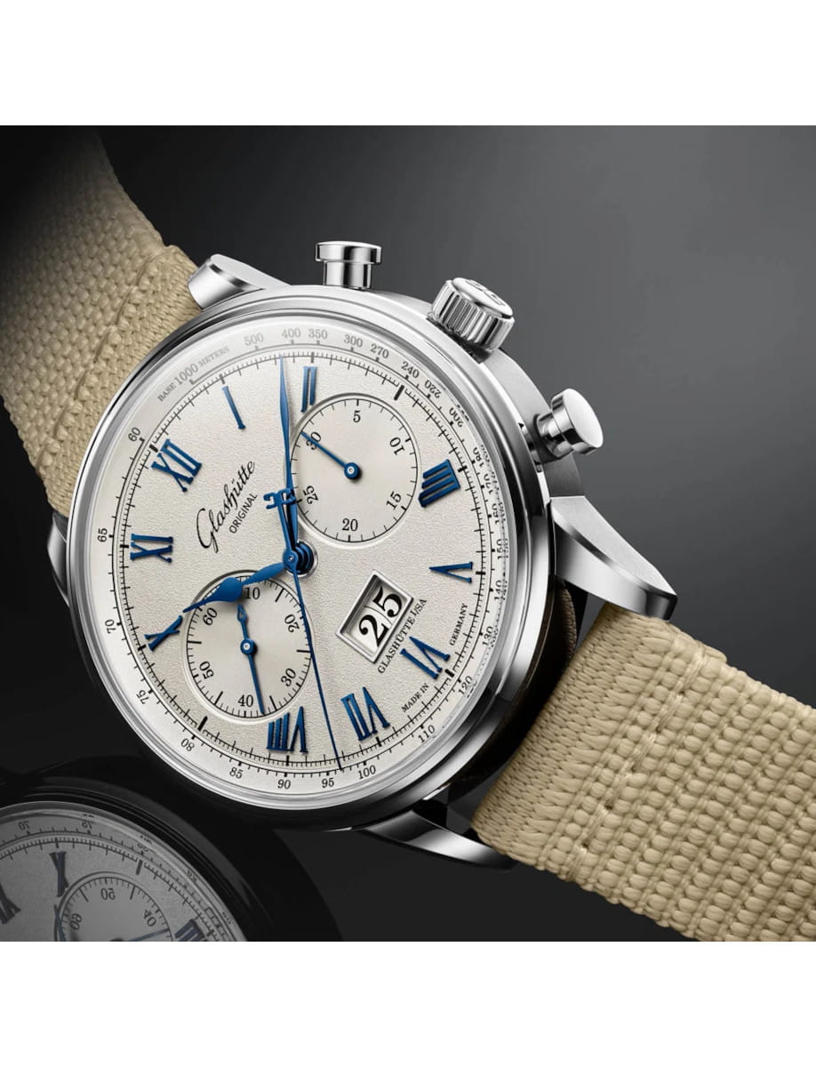 Stainless steel wristwatch with a beige fabric strap, white dial, blue Roman numerals, and multiple subdials (chronograph). See date window at 4 o'clock.