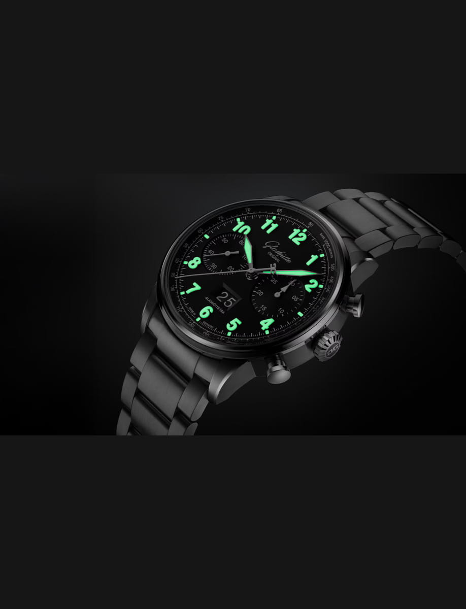 Close-up of a black stainless steel wristwatch with luminous green numerals and hands on a dark background.