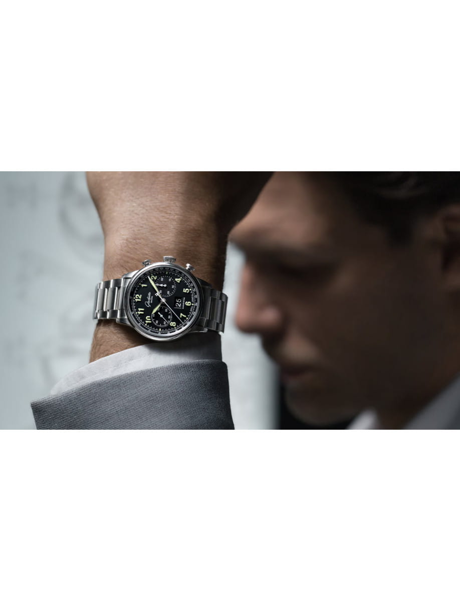 Close-up of a man's left wrist wearing a silver metal watch with a black dial and white numerals; blurred profile in the background