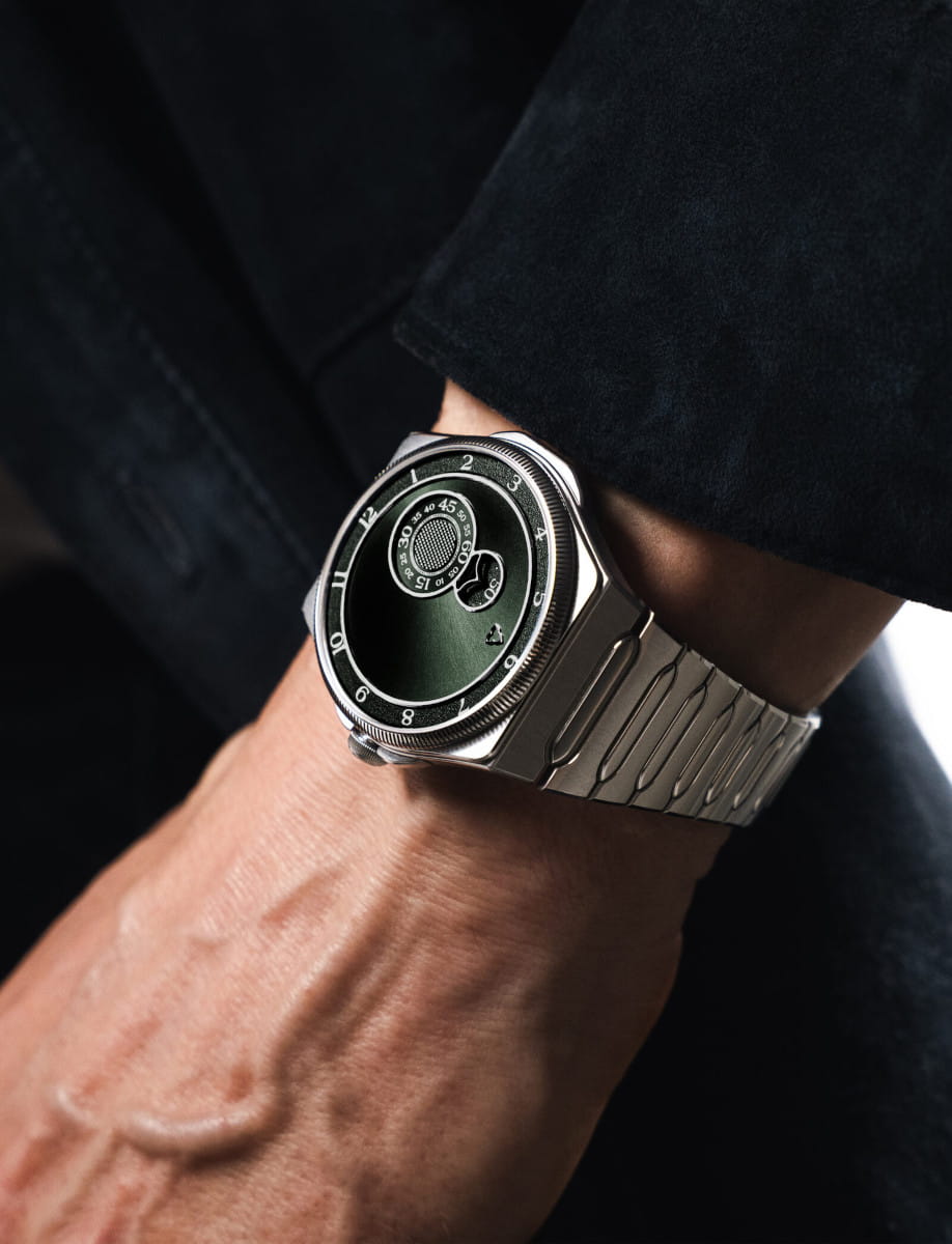 Close-up of a silver metal wristwatch on a dark sleeve, featuring a black circular dial and numbered bezel.