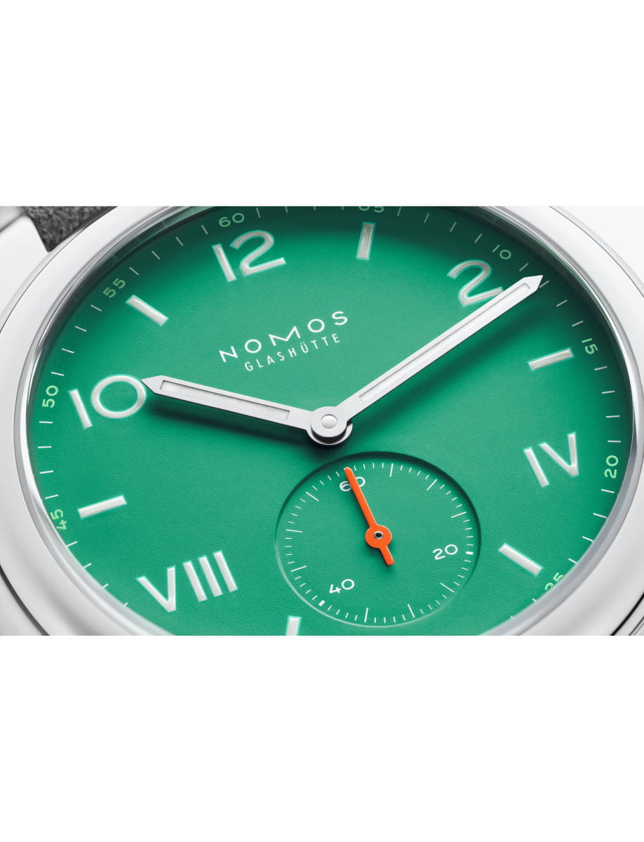 Close-up of a green NOMOS Glashütte wristwatch dial with silver hands and an orange seconds hand, featuring Roman numerals and a small subdial.