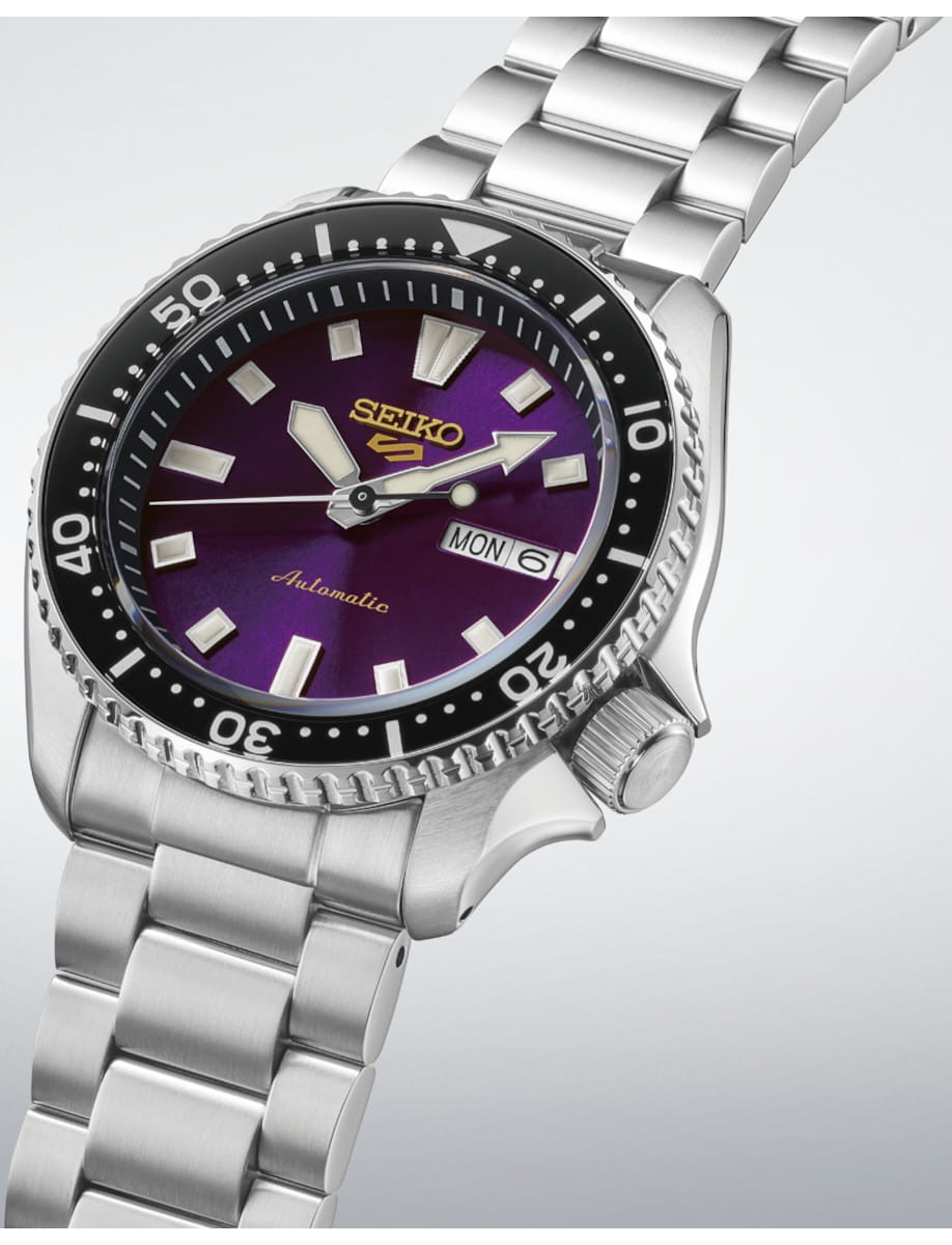 Close-up of a Seiko automatic wristwatch with a purple dial, black bezel, and stainless steel bracelet, showing day/date at 3 o'clock.