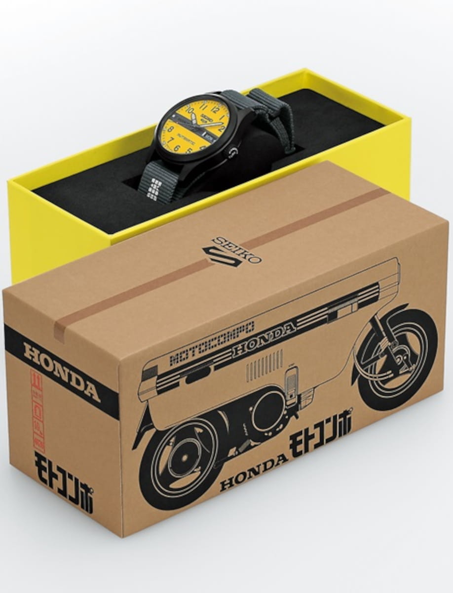 Wristwatch with a yellow dial in a black case on a yellow display box, resting on a Honda motorcycle-themed cardboard box.